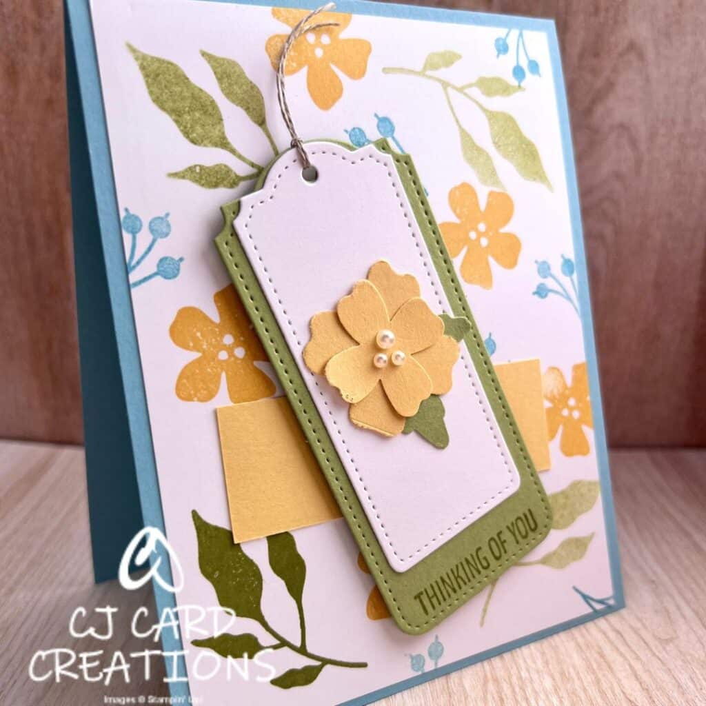 blue card with white layer stamped with green leaves, blue berries, and yellow flowers, with a green tag reading "thinking of you" and a white tag with a 3D yellow flower with pearls