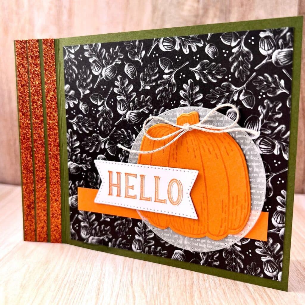 Green card with glittery orange strips, a black and white background of leaves and acorns, an orange pumpkin, and "hello" in orange ink