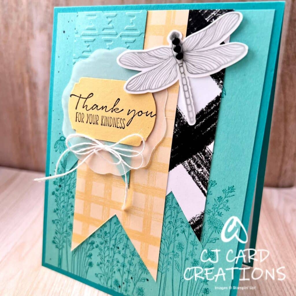 An aqua card with stamped flowers, a light aqua gingham-embossed banner, a yellow plaid banner, and a black and white plaid banner, finished with a dragonfly and a label that says "Thank you for your kindness"
